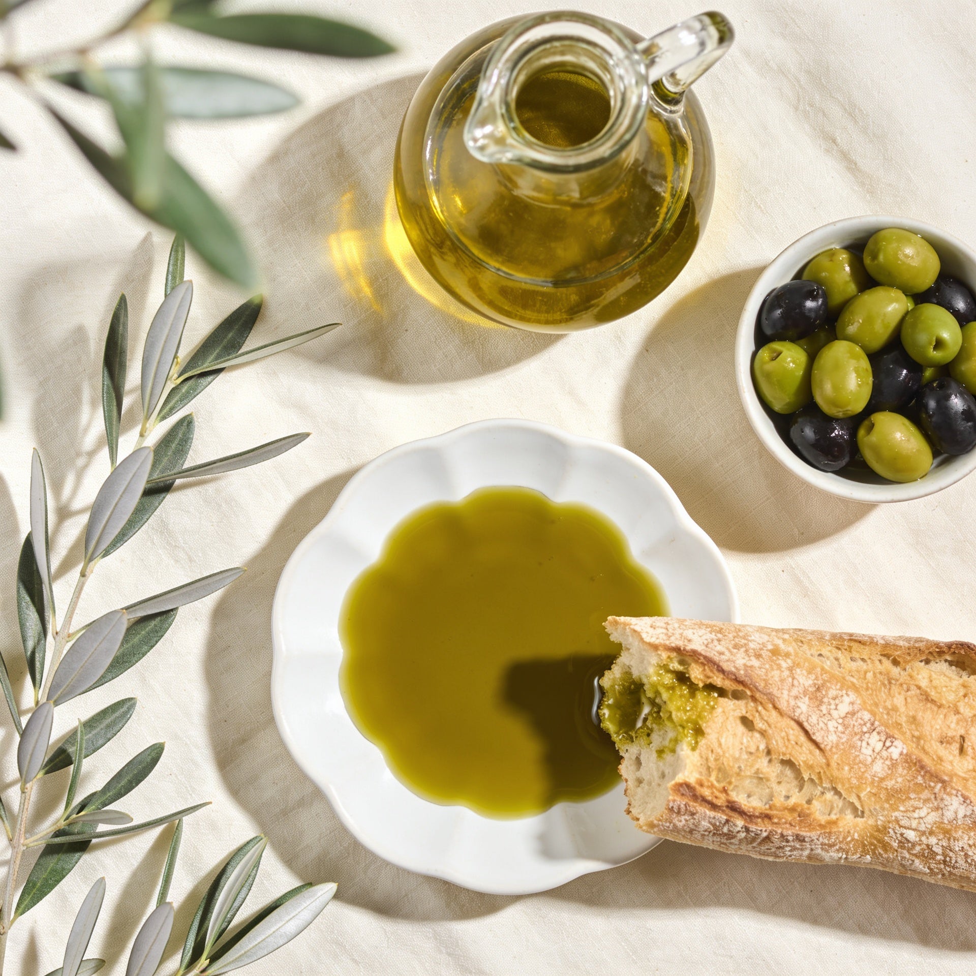 Olivenöl in weißer Schale mit Oliven, Brot und Olivenzweigen auf hellem Leinen